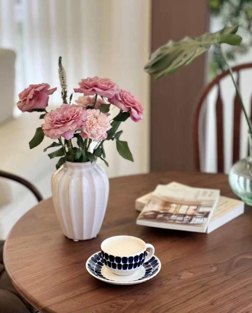 Custom porcelain vase from China with pink roses and carnations on a wooden table next to a patterned coffee cup and design books